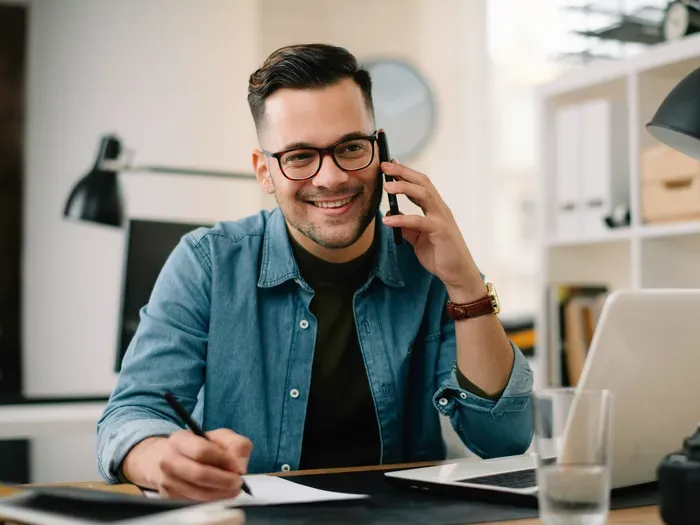Professional man smiling while talking on the phone and taking notes at his desk with a laptop nearby for Town Network Hub
